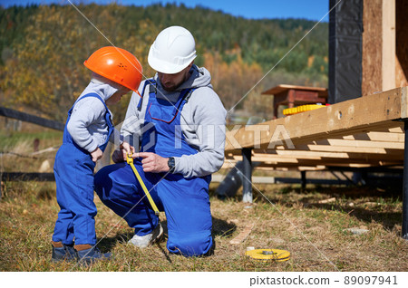 Father with toddler son building wooden frame house. Male builder and kid playing with tape measure on construction site, wearing helmet and blue overalls on sunny day. Carpentry and family concept. 89097941