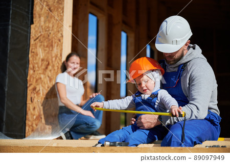 Father, mother and son building wooden frame house. Toddler boy helping his daddy, while woman looking for them on construction site. Guys wearing helmet and blue overalls. Carpentry, family concept. 89097949