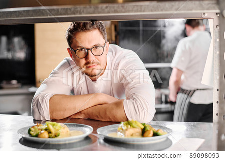 Portrait of young professional cook in eyeglasses looking at camera behind the counter with ready dishes Portrait of young professional cook in eyeglasses looking at camera behind the counter with ready dishes 89098093