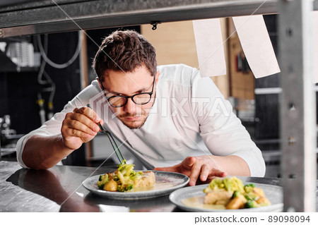 Young serious chef in eyeglasses decorating main dish on plate on counter before delivery 89098094