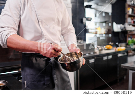 Close-up of chef in apron whipping eggs in bowl with a whisk while cooking in kitchen Close-up of chef in apron whipping eggs in bowl with a whisk while cooking in kitchen 89098119
