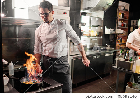 Serious young chef in uniform cooking vegetables on pan in flambe style on cooker in commercial kitchen 89098140