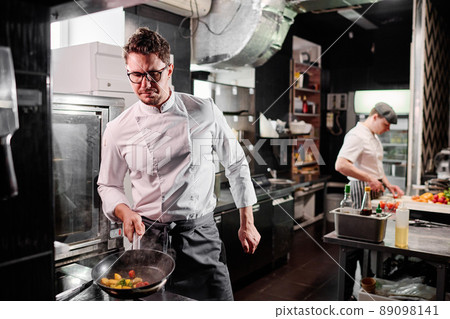 Professional chef in apron using cooker to fry vegetables on pan during work in kitchen with colleague in background 89098141
