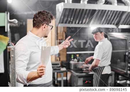 Chef gesturing his assistant to prepare food for two people during their work in commercial kitchen 89098145