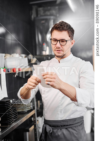 Professional cook in apron holding paper in his hands and examining order of guest before cooking 89098146
