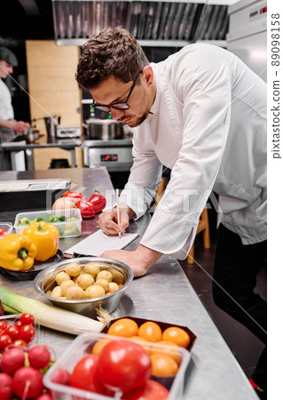 Young chef making a menu at table with food ingredients during his work in kitchen of restaurant 89098158