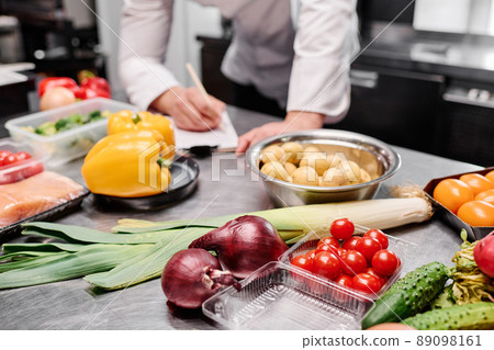 Close-up of fresh vegetables on table in commercial kitchen with chef making menu in background 89098161