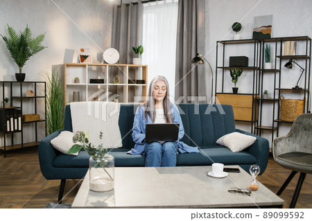 Happy smiling senior woman, sitting on blue sofa and studying with laptop at home 89099592