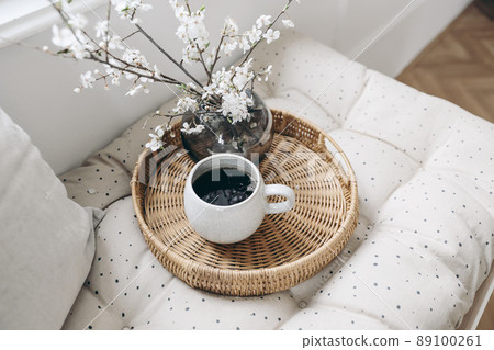 Spring breakfast still life composition. Cup of coffee and wickered tray on French mattress. Feminine styled photo. Floral scene with blurred white cherry tree blossoms. Bench near window. Selective Spring breakfast still life composition. Cup of coffee and wickered tray on French mattress. Feminine styled photo. Floral scene with blurred white cherry tree blossoms. Bench near window. Selective 89100261