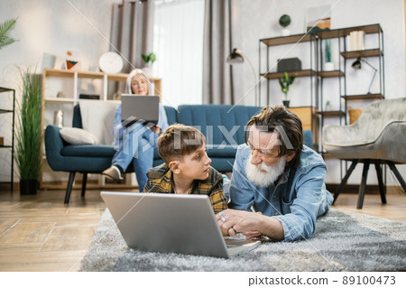 Grandmother relaxing using laptop on sofa while her grandchild playing on laptop with grandpa. Grandmother relaxing using laptop on sofa while her grandchild playing on laptop with grandpa. 89100473