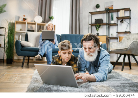 Grandmother relaxing using laptop on sofa while her grandchild playing on laptop with grandpa. Grandmother relaxing using laptop on sofa while her grandchild playing on laptop with grandpa. 89100474