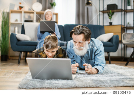 Grandmother relaxing using laptop on sofa while her grandchild playing on laptop with grandpa. Grandmother relaxing using laptop on sofa while her grandchild playing on laptop with grandpa. 89100476