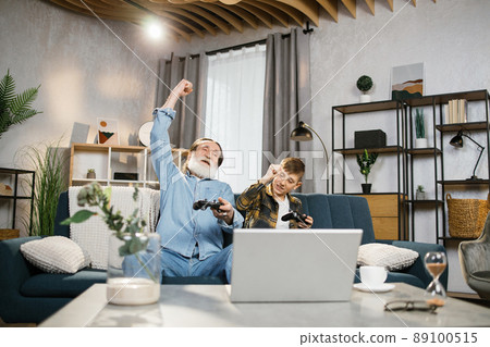 Elder man and his grandson sitting on couch and using wireless joysticks for playing video games 89100515
