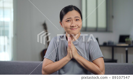 Asian woman holding hands in prayer, looking at camera sitting on sofa at home. 89107547