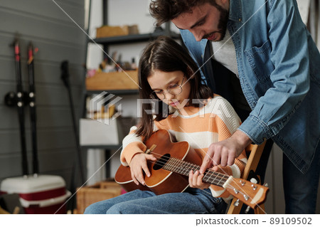 Little girl in eyeglasses learning to play guitar while father bending over her 89109502