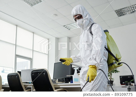 Black man in hazmat suit spraying floor of openspace office with disinfectant 89109775