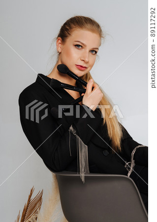 Beautiful middle aged woman makeup waving one makeup brush, winking at the camera and smiling. Blond hair and a black jacket on a light background. Beautiful middle aged woman makeup waving one makeup brush, winking at the camera and smiling. Blond hair and a black jacket on a light background. 89117292
