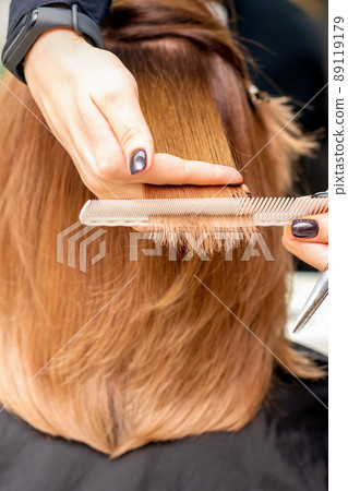 Hands of hairdresser hold hair strand between his fingers making haircut of long hair of the young woman with comb and scissors in hairdresser salon, close up Hands of hairdresser hold hair strand between his fingers making haircut of long hair of the young woman with comb and scissors in hairdresser salon, close up 89119179