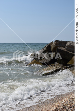 The waves of the turquoise sea break on the coastal boulders and a dense white foam is formed. Clear blue sky. White waves breaking on sandy beach through gap in rocks on sea The waves of the turquoise sea break on the coastal boulders and a dense white foam is formed. Clear blue sky. White waves breaking on sandy beach through gap in rocks on sea 89120298