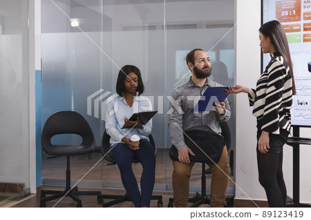 Businesswoman giving cv resume to man applicant asking about work experience during interview meeting in startup company office. Diverse candidates waiting for career opportunity in lobby area 89123419