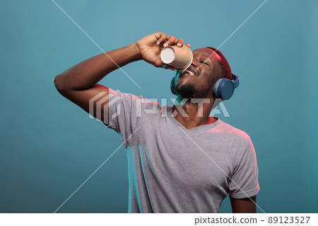 Excited man drinking coffee from cup and wearing headphones to listen to radio music on audio gadget. Casual person with wireless earphones enjoying drink and mp3 sound to relax. Excited man drinking coffee from cup and wearing headphones to listen to radio music on audio gadget. Casual person with wireless earphones enjoying drink and mp3 sound to relax. 89123527