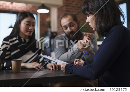 Young adult recruiter sitting at desk in office space while reviewing job applicants CVs. Multiethnic candidates talking with startup company interviewer while talking about career oportunities. 89123575