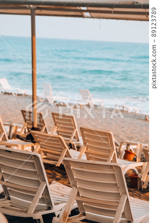 Empty Sun loungers in front of sea. Sun chairs on sandy beach. Big waves stormy weather. An empty chaise longue and folded umbrella stand at the edge of the sea Empty Sun loungers in front of sea. Sun chairs on sandy beach. Big waves stormy weather. An empty chaise longue and folded umbrella stand at the edge of the sea 89123789