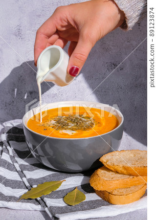 Woman adding cream to Homemade carrot ginger curcuma soup. Female hand pouring milk into Seasonal Pumpkin traditional soup with creamy silky texture. Healthy vegan clean eating. 89123874