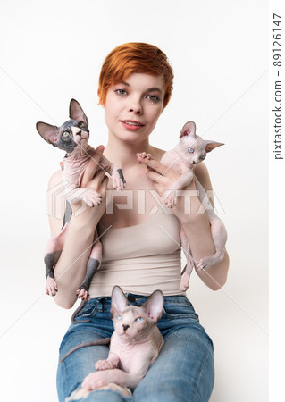 Redhead young woman holding two Sphynx Cat in hands and one kitten lying on her legs. Pretty woman with short hair in T-shirt and jeans looking camera, lying down on white background. Part of series 89126147