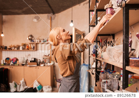 Female artist putting jar at the shelves while preparing drawing at her art space 89126678