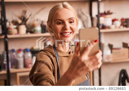Cheerful female artist holding mobile phone and making self portrait on it at her art studio Cheerful female artist holding mobile phone and making self portrait on it at her art studio 89126681