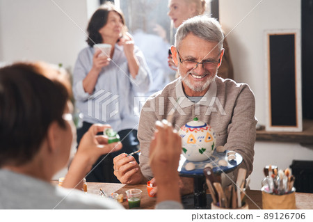 Two pensioners laughing while sitting at the table and decorating hand made teapots 89126706