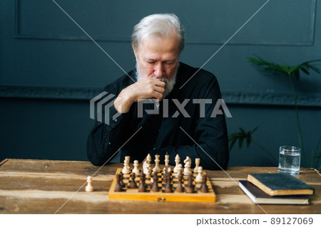 Front view of thoughtful bearded mature pensioner male thinking about chess move sitting at home and playing chess alone. Pensive senior adult man thinking game strategy at table with chessboard. 89127069