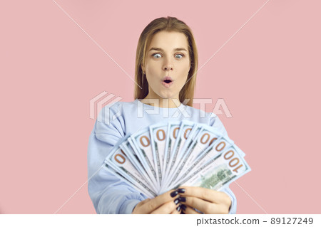 Amazed young woman with wide open mouth and eyes looking at dollar bills holding in hands. 89127249