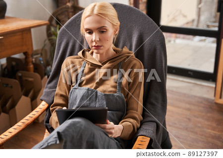 Artist woman sitting at the armchair and looking at her tablet with serious face Artist woman sitting at the armchair and looking at her tablet with serious face 89127397