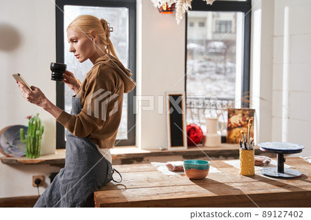 Female artist holding cup of tea and looking at her smartphone while relaxing before the work 89127402