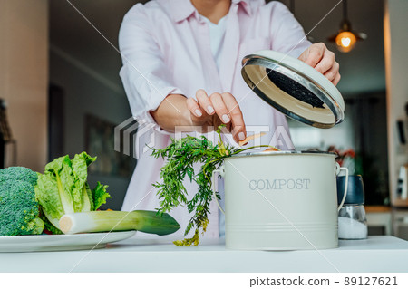 Compost the kitchen waste, recycling at home. Woman putting vegetables cutted leftovers into the garbage, compost bin on the table of kitchen. Environmentally responsible, ecology. Selective focus. 89127621