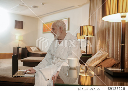 A gray-haired man in a white robe watching something on a laptop 89128226