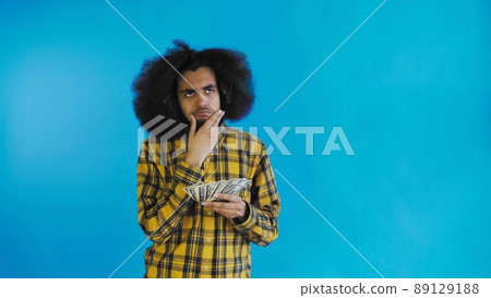 A happy African-American holding money in his hands and thinking about what to spend it on, stands isolated on a blue background A happy African-American holding money in his hands and thinking about what to spend it on, stands isolated on a blue background 89129188