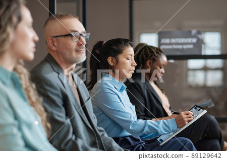 Young serious businesswoman filling resume form for job interview while sitting in waiting room with people Young serious businesswoman filling resume form for job interview while sitting in waiting room with people 89129642
