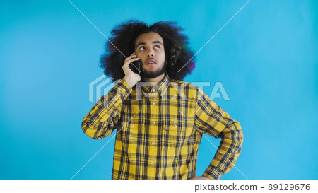 A young man with an African hairstyle on a blue background is talking on the phone. On a colored background 89129676