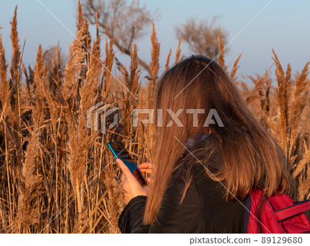 Pretty young woman in black coat with backpack smartphone on pampas grass sunset sky dry reeds Millennial long hair girl Pretty young woman in black coat with backpack smartphone on pampas grass sunset sky dry reeds Millennial long hair girl 89129680
