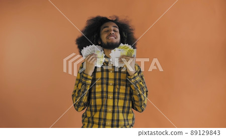 A happy African-American man holding a fan of banknotes in his hands and looking at the camera, standing isolated on an orange background 89129843