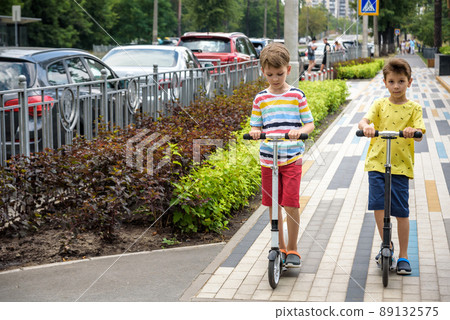 Two attractive European boys brothers, wearing red and white checkered shirts, standing on scooters in the park. They laughing, smiling, hugging and having fun. Active leisure time with kids 89132575