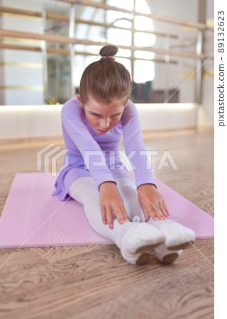 Beautiful little ballerina in a lilac leotard and white tights doing stretching exercises on a pink karemat in dance studio. Beautiful little ballerina in a lilac leotard and white tights doing stretching exercises on a pink karemat in dance studio. 89132623