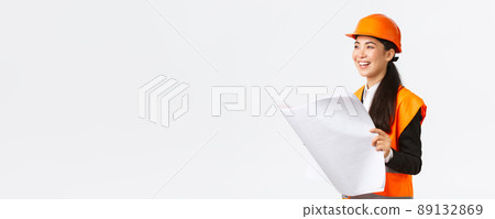 Satisfied happy asian female chief engineer looking at results of construction works, studying blueprint with pleased face, wearing safety helmet and reflective clothings at building zone 89132869
