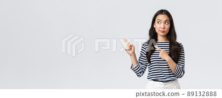 Lifestyle, beauty and fashion, people emotions concept. Curious and interested cute asian girl peeking and pointing fingers left at new promo banner with special discount in shop 89132888