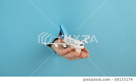 A female hand sticking out of a hole from a blue background holds a model of an airplane. A female hand sticking out of a hole from a blue background holds a model of an airplane. 89133174