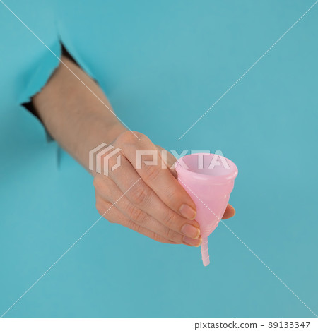 A female hand sticking out of a hole from a blue background holds a pink menstrual cup. A female hand sticking out of a hole from a blue background holds a pink menstrual cup. 89133347