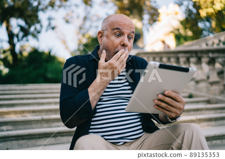 Caucasian senior 60s with shock expression on face reading received news on digital tablet Caucasian senior 60s with shock expression on face reading received news on digital tablet 89135353